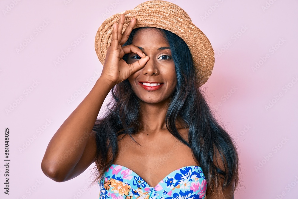 Young indian girl wearing bikini and summer hat smiling happy doing ok sign with hand on eye looking through fingers Young indian girl wearing bikini and summer hat smiling happy doing ok sign with hand on eye looking through fingers