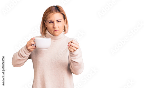 Young blonde woman holding coffee annoyed and frustrated shouting with anger, yelling crazy with anger and hand raised