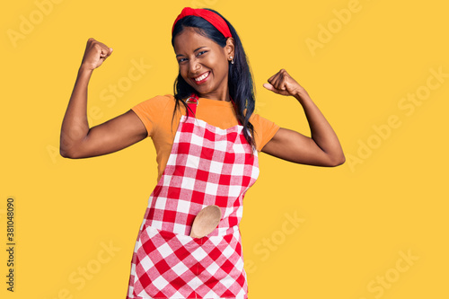 Tela Young indian girl wearing professional baker apron showing arms muscles smiling proud