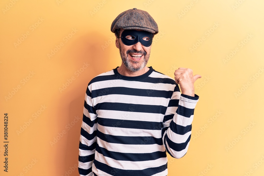 Middle age handsome burglar man wearing cap and mask over isolated yellow background pointing thumb up to the side smiling happy with open mouth Middle age handsome burglar man wearing cap and mask over isolated yellow background pointing thumb up to the side smiling happy with open mouth