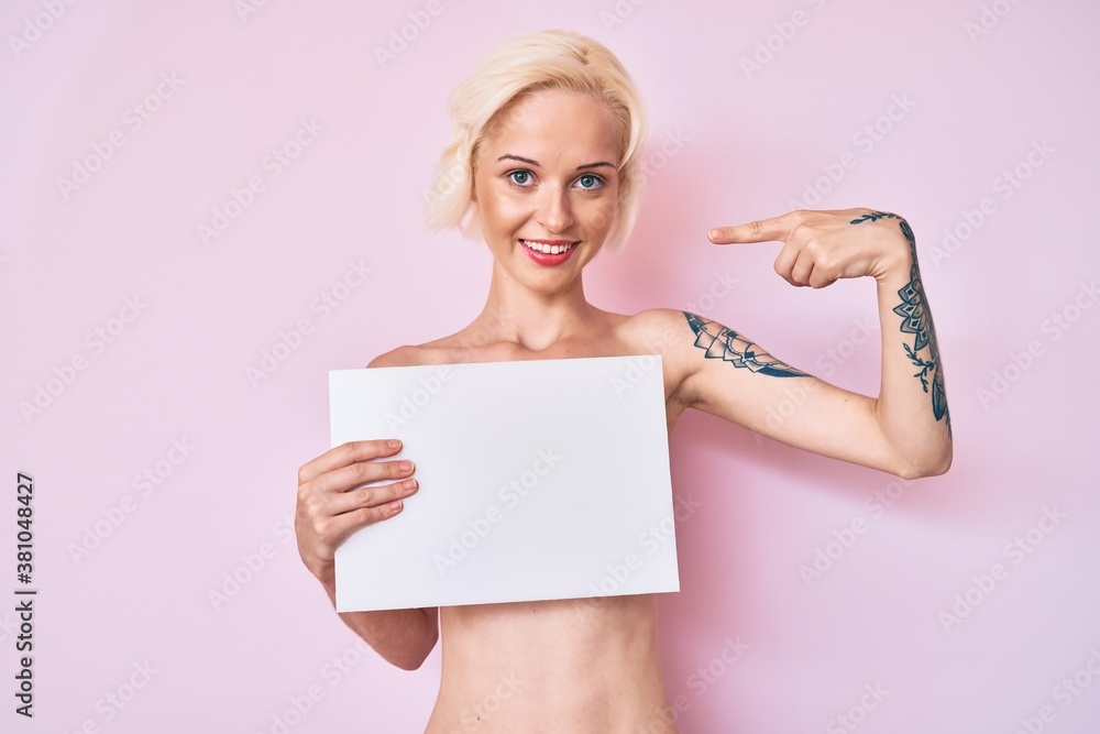 Young blonde woman with tattoo standing shirtless holding empty blank banner pointing finger to one self smiling happy and proud Young blonde woman with tattoo standing shirtless holding empty blank banner pointing finger to one self smiling happy and proud