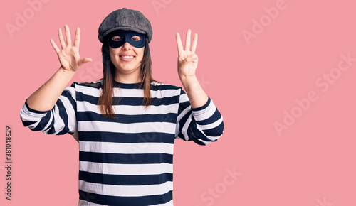 Young beautiful brunette woman wearing burglar mask showing and pointing up with fingers number eight while smiling confident and happy.