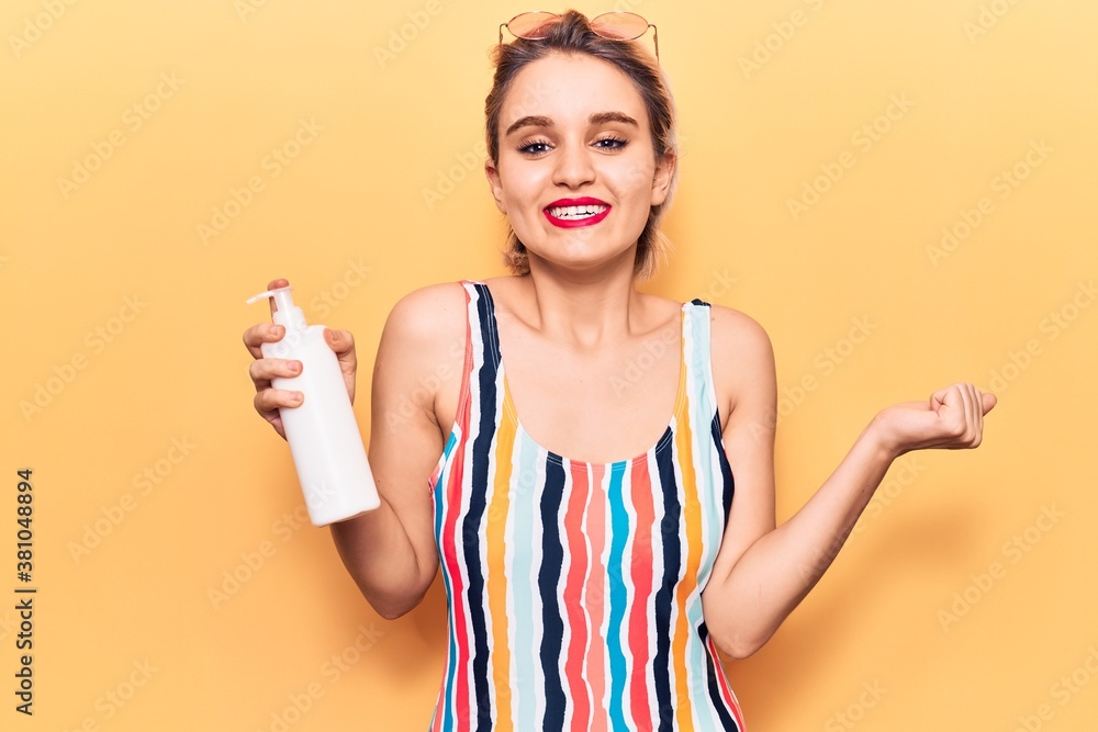 Young beautiful blonde woman wearing swimwear holding sunscreen lotion screaming proud, celebrating victory and success very excited with raised arm Young beautiful blonde woman wearing swimwear holding sunscreen lotion screaming proud, celebrating victory and success very excited with raised arm