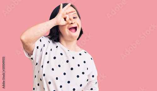 Brunette woman with down syndrome wearing casual clothes doing ok gesture shocked with surprised face, eye looking through fingers. unbelieving expression.