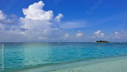 Fototapeta Naklejka Na Ścianę i Meble -  White whimsical clouds are above the calm aquamarine ocean. There is no one on the sandy beach. In the distance you can see a path above the water, an island. There is a boat on the horizon. Maldives.
