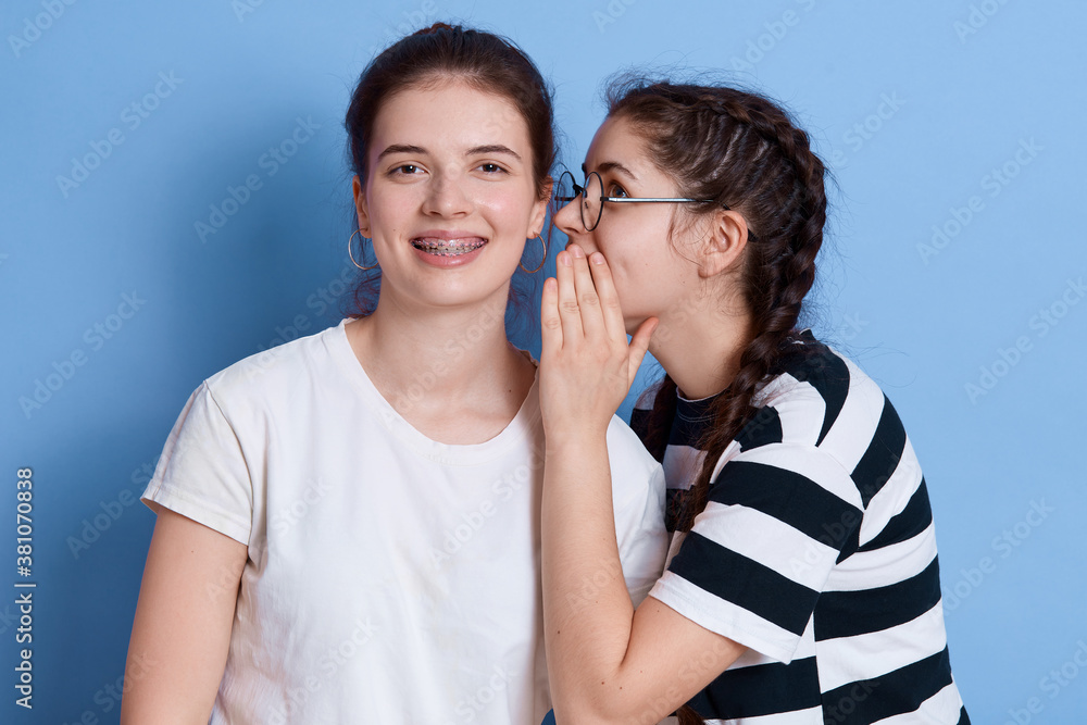 Two excited young girls dressed in summer clothes gossiping isolated over blue background, lady with glasses whispering something to ear, girls expressing happiness. Two excited young girls dressed in summer clothes gossiping isolated over blue background, lady with glasses whispering something to ear, girls expressing happiness.