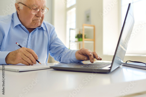 Senior man in glasses using laptop and taking notes in notebook sitting at desk at home