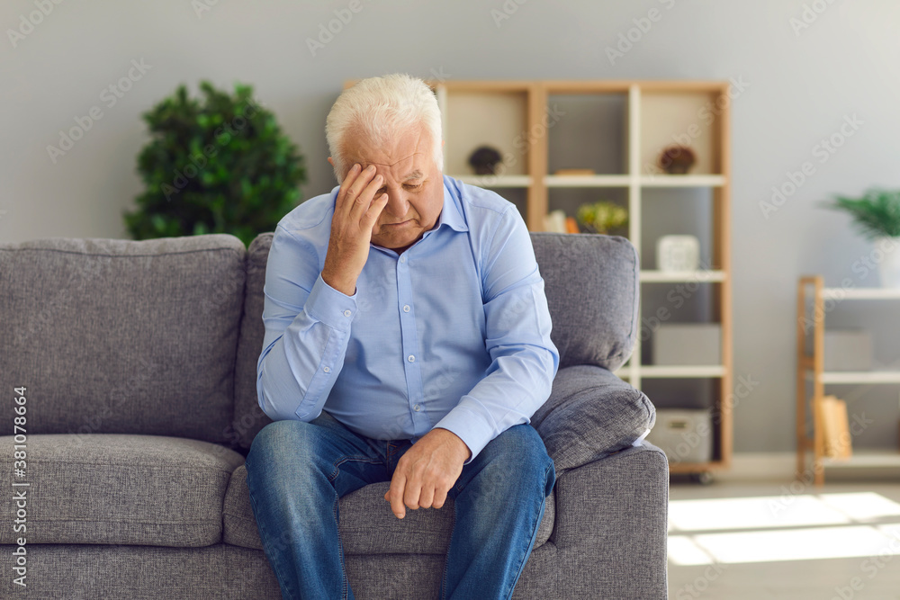 Depressed senior man sitting on sofa at home suffering from loneliness or bad headache