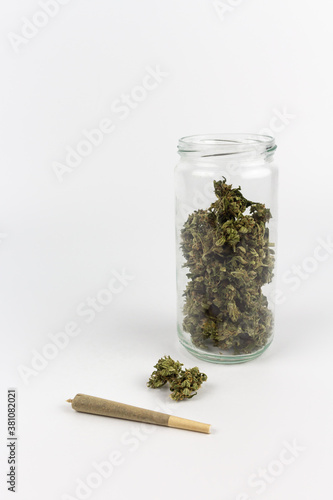 View of a glass jar with marijuana buds inside and a bud with a joint on a white background 