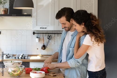 Happy Caucasian couple tenants enjoy lazy morning in modern kitchen cooking together. Smiling loving young man and woman renters preparing healthy delicious vegetarian food at new own home.