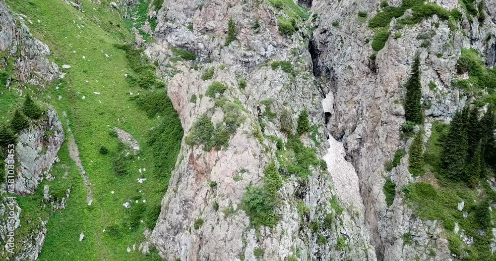 Top view of steep mountain cliffs and an ice cave. An avalanche descended into the gorge, and a snow tunnel (ice grotto) was formed. Mountainous terrain, spruce trees and green grass grow. Top view of steep mountain cliffs and an ice cave. An avalanche descended into the gorge, and a snow tunnel (ice grotto) was formed. Mountainous terrain, spruce trees and green grass grow.