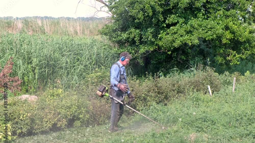 Senior man mows grass with petrol brush cutter. Man wearing overalls, protective goggles, soundproof headphones and work gloves. Full-length side view Senior man mows grass with petrol brush cutter. Man wearing overalls, protective goggles, soundproof headphones and work gloves. Full-length side view