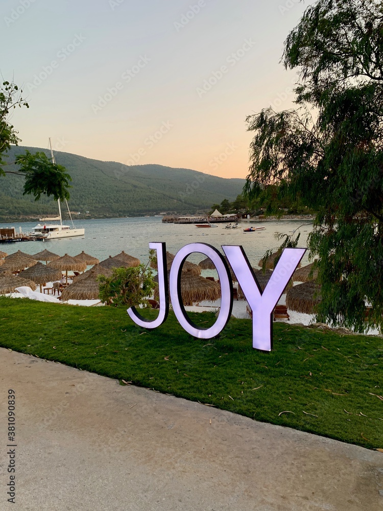 Fototapeta premium Large letters: JOY, Panoramic view of the emerald Aegean coast with white sand, bamboo palms and sun loungers. Luxury leisure relaxation concept. White beach with a mountain in the background
