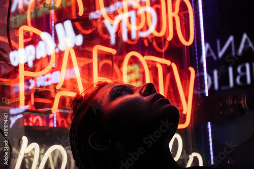 A hooligan girl stands against the background of a neon sign in the evening in the city.