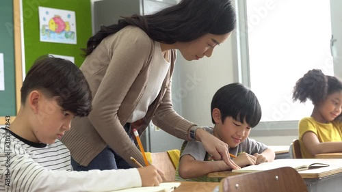 Asian school teacher assisting students in classroom. Young woman working in school helping boy with his writing, education, support, care.