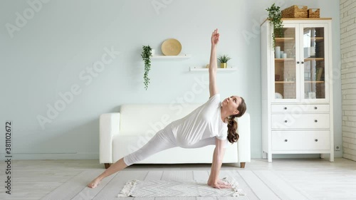 Flexible brunette pregnant woman does utthita parsvakonasana practicing yoga position on floor against designer furniture in spacious room at home photo