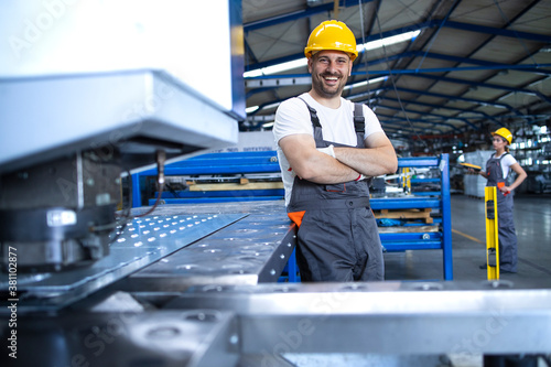 Portrait of factory worker in protective uniform and hardhat standing by industrial machine at production line. People working in industry.