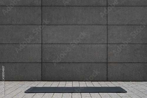 empty and blank foldable black yoga mat on grey sidewalk near empty street outdoor in front of grungy concrete textured wall with copy space is a perfect backdrop for text and yoga fitness graphics