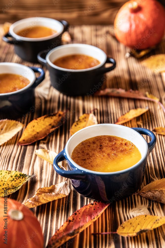 orange Creme Brulee aus Kürbis vom Hokkaido mit karamellisierter Zuckerkruste in Förmchen auf einem braunen Holztisch mit Laubblättern im Herbst orange Creme Brulee aus Kürbis vom Hokkaido mit karamellisierter Zuckerkruste in Förmchen auf einem braunen Holztisch mit Laubblättern im Herbst