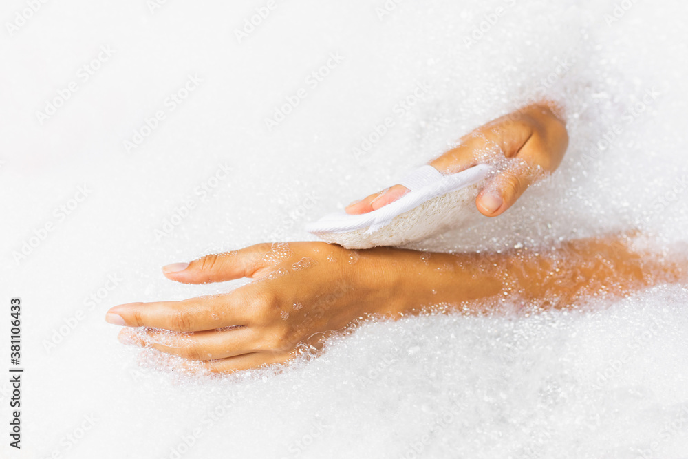 Fototapeta premium Woman washing hands in bath tub with foam bubbles and use natural jute sponge. Hands close-up. Spa treatment, body care.