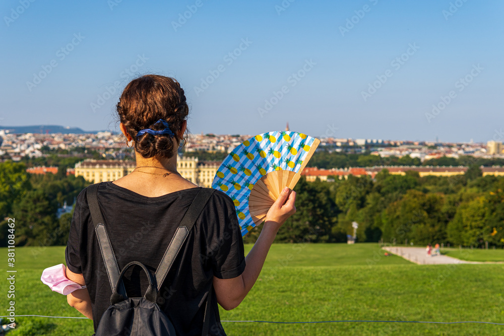 Young woman standing with here back to the camera wearing a black backpack and holding a hand fan on a green hill and looking to the horizon view of the city vienna 