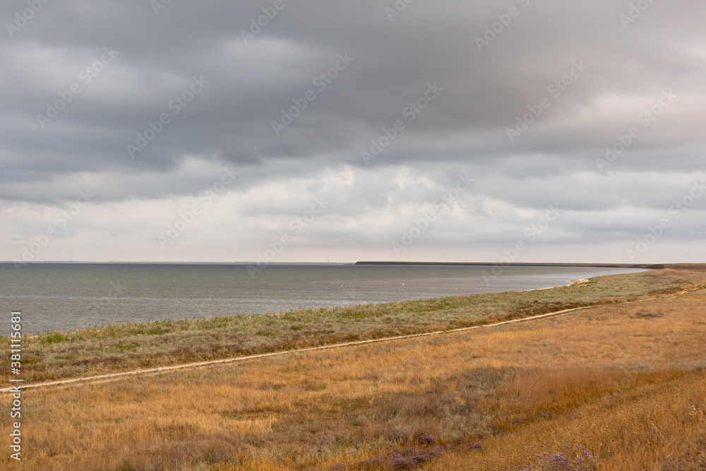 Grey high layered grey epic clouds above wild autumn yellow dry field with lake water line. Heaven cloudscape air view