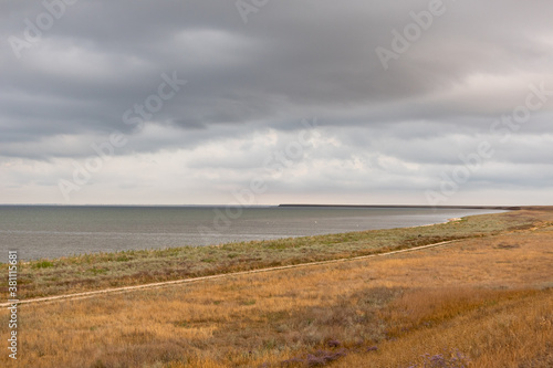 Grey high layered grey epic clouds above wild autumn yellow dry field with lake water line. Heaven cloudscape air view