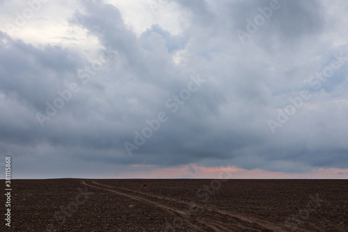 Autumn cultivated land rural bare field with dirt road going to horizon with dramatic sunrise colored gorgeous clouds. Plowed field with grey epic sunset layered cloudscape