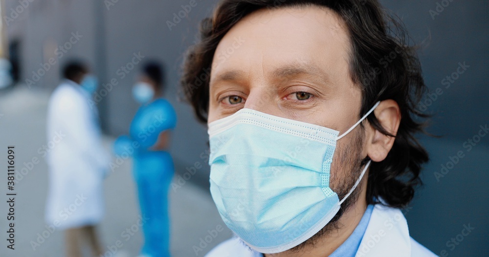 Portrait of Caucasian handsome man doctor in medical mask looking at camera. Close up of male physician in respiratory protection. African American medics colleagues on background.