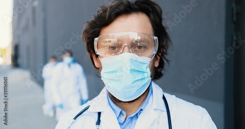 Portrait of Caucasian good-looking man doctor in medical mask, goggles and with stethoscope looking at camera. Close up male physician in respiratory protection. Multi ethnic doctors on background.