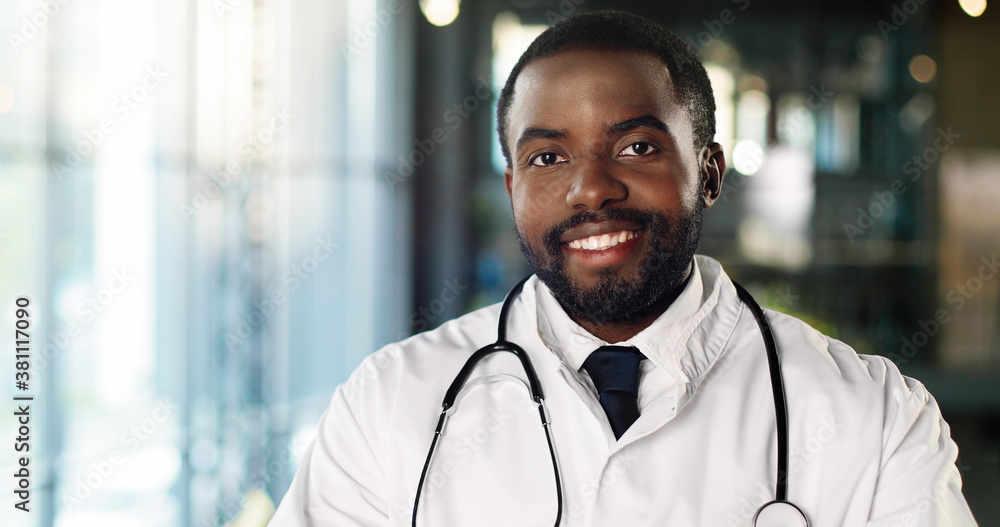 Portrait of African American young man doctor with stethoscope looking at camera and smiling cheerfully. Handsome male physician smile. Medic in white gown. Indoors. Portrait of African American young man doctor with stethoscope looking at camera and smiling cheerfully. Handsome male physician smile. Medic in white gown. Indoors.