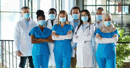 Mixed-races busy team of male and female doctors posing to camera and crossing hands in hospital. International group of medics in medical masks. Protected multi ethnic physicians and nurses in clinic