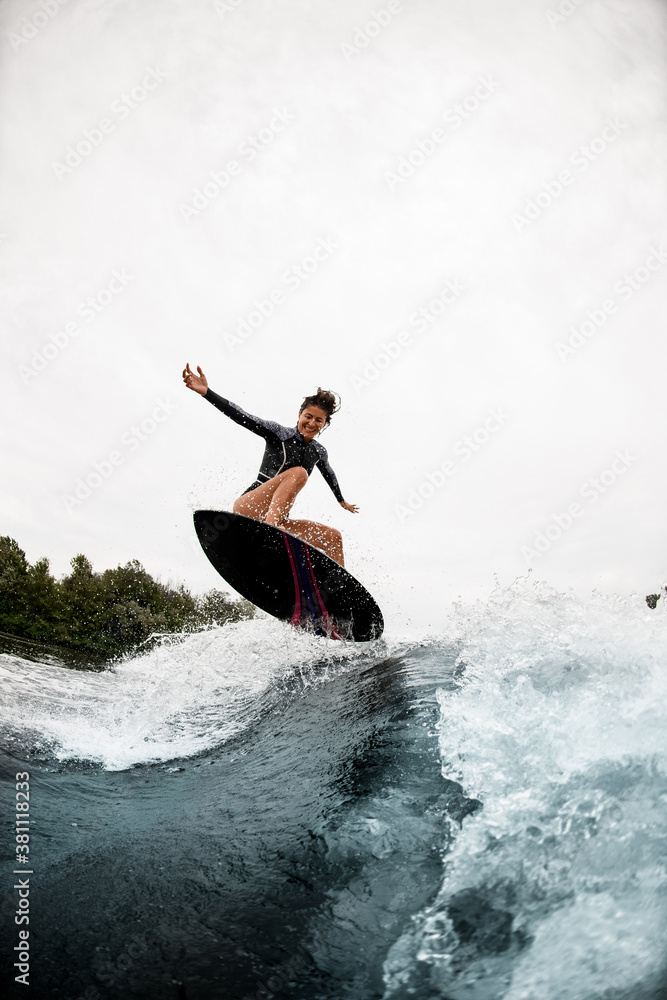 cheerful woman in black wetsuit effectively jumps on surfboard on wave