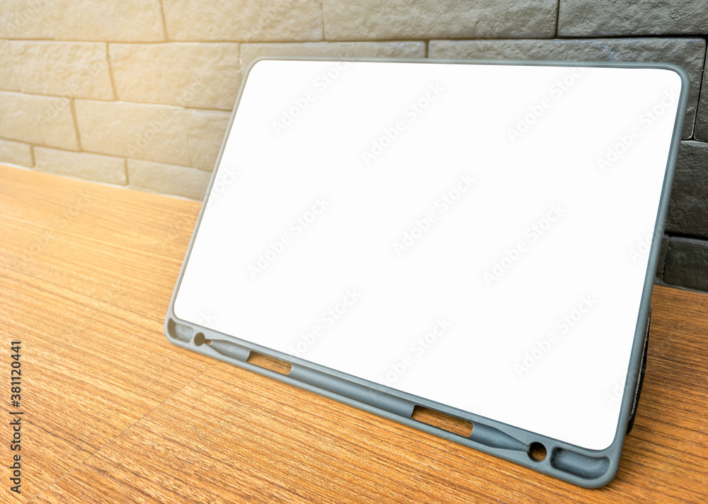 Blank space or empty space of tablet screen, white colour. Tablet on wooden desk, standing by its cover protection, grey colour brick wall background, perspective view, warm light tone. Blank space or empty space of tablet screen, white colour. Tablet on wooden desk, standing by its cover protection, grey colour brick wall background, perspective view, warm light tone.