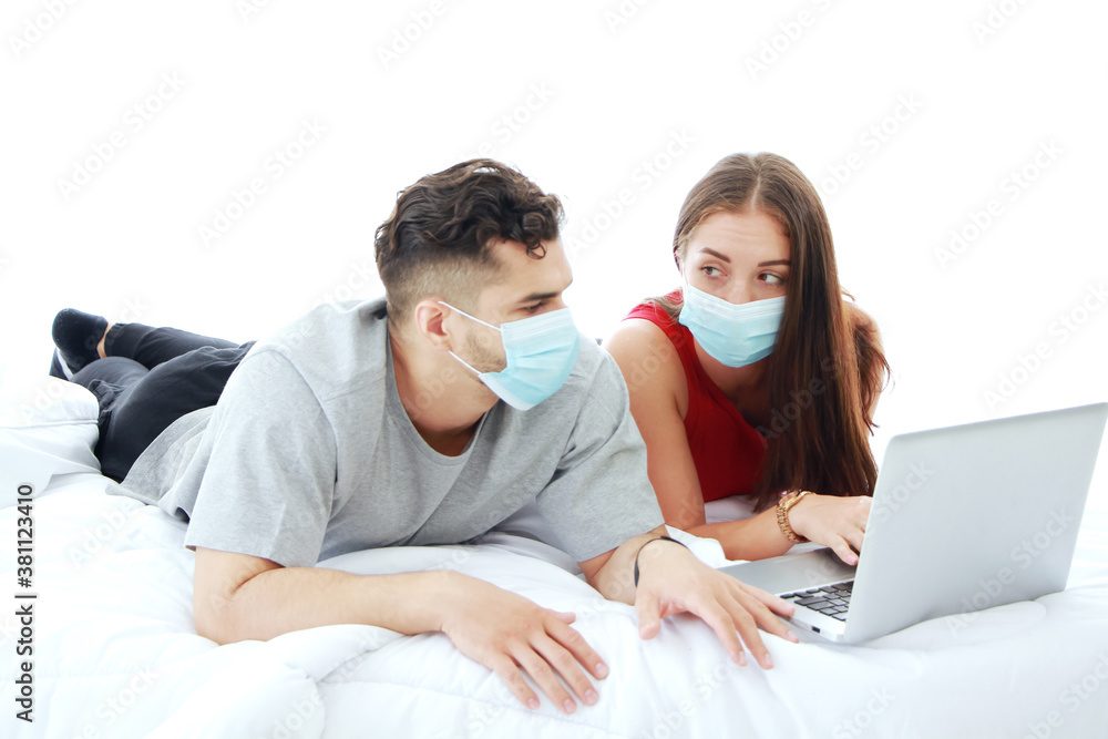 Caucasian couple wearing mask prevent covid-19 virus is laying with laptop in social media technology in bedroom. Social distancing and healthcare for Married family lifestyle on holiday Concept.