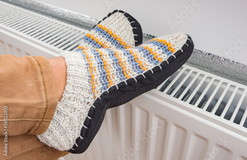 A woman is dressed in warm knitted woolen slippers  warm her legs on a heating radiator. The symbolic image of the heating season at home. photo