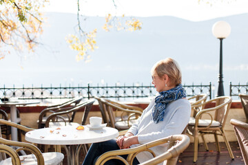 Happy senior woman in cozy autumn cafe outdoors. Retired stylish lady drinking morning coffee and enjoying fall season by sea. Concept of wellbeing, Indian summer, life satisfaction. Lifestyle moment.