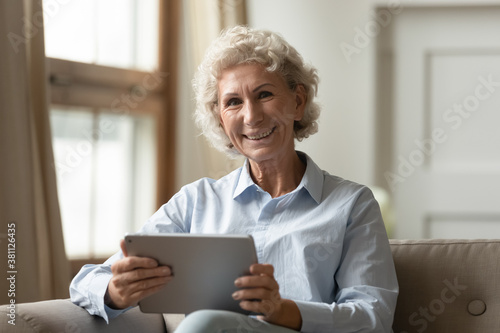 Easy to work with. Happy retired 60s female posing for portrait on cozy couch at home looking at camera with digital pad gadget, showing it is simple and convenient to call, use app or surf internet