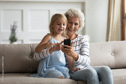 Look here, my darling. Senior grandma and small girl grandkid spending time together using phone, texting message, chatting, dialing number, watching funny photos in social network, browsing internet