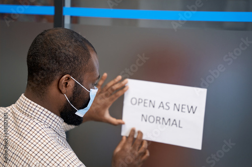Rear view of young african man wearing protective face mask sticking open sign with text OPEN AS NEW NORMAL on the glass door in the office