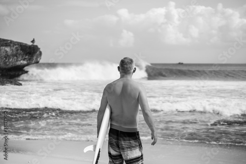 Black and white portrait of handsome shirtless man surfer , holding white surf board and cliff, rocks , wave on background