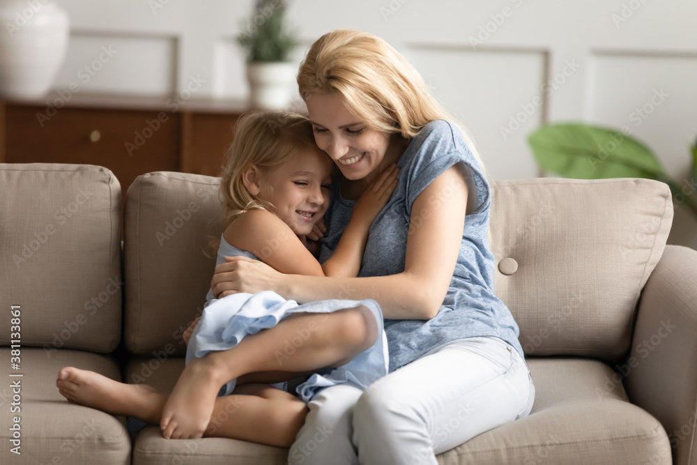 Warm embraces at home. Little daughter and young mommy hugging on couch, happy stepmom and beloved girl child in care snuggling down on sofa at living room feeling love in cuddles of one another