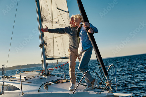 Elegant senior man pointing at the horizon while standing with his wife on the side of a sail boat or yacht deck floating in a calm blue sea, enjoying amazing view