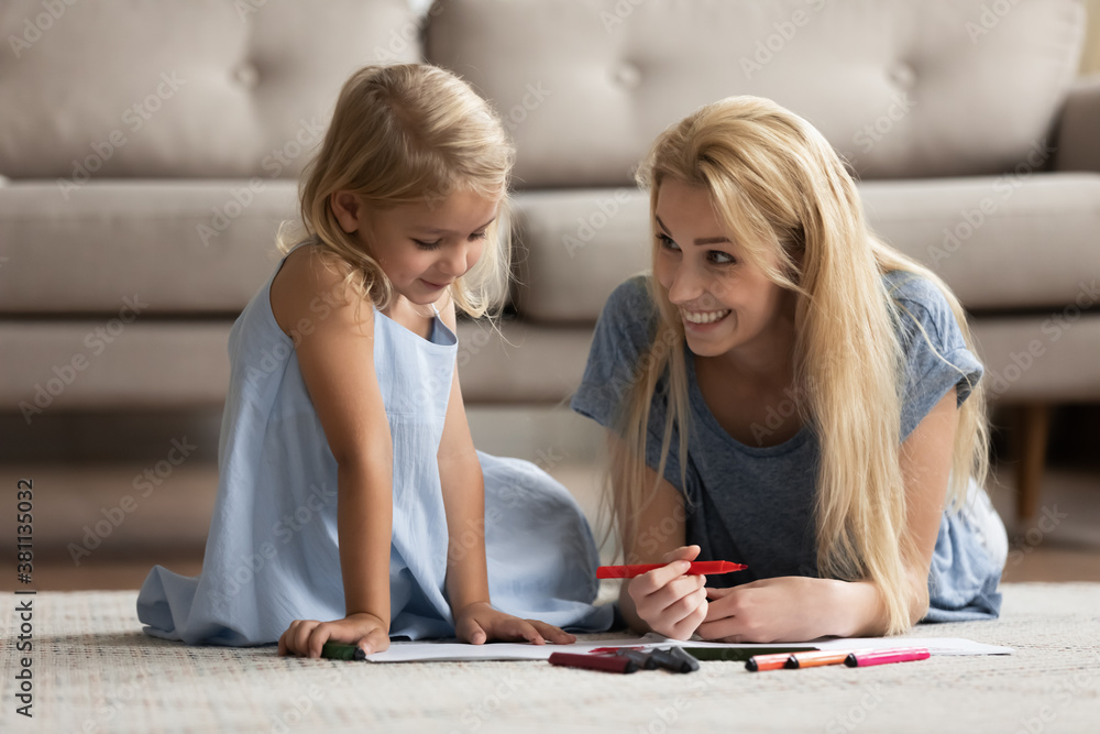 Fototapeta premium Creative activity. Happy smiling young mother spending leisure time with little kid daughter lying on heated floor in living room drawing funny pictures with bright colorful marker pens in paper album
