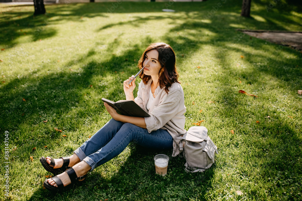 Young woman student sitting on grass in a park with notebook, thinking. Young woman student sitting on grass in a park with notebook, thinking.