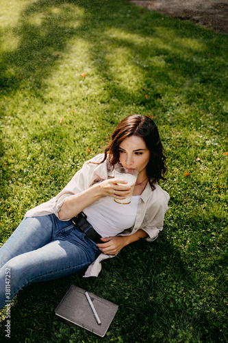 Young woman drinking coffee, sitting on green grass in park.