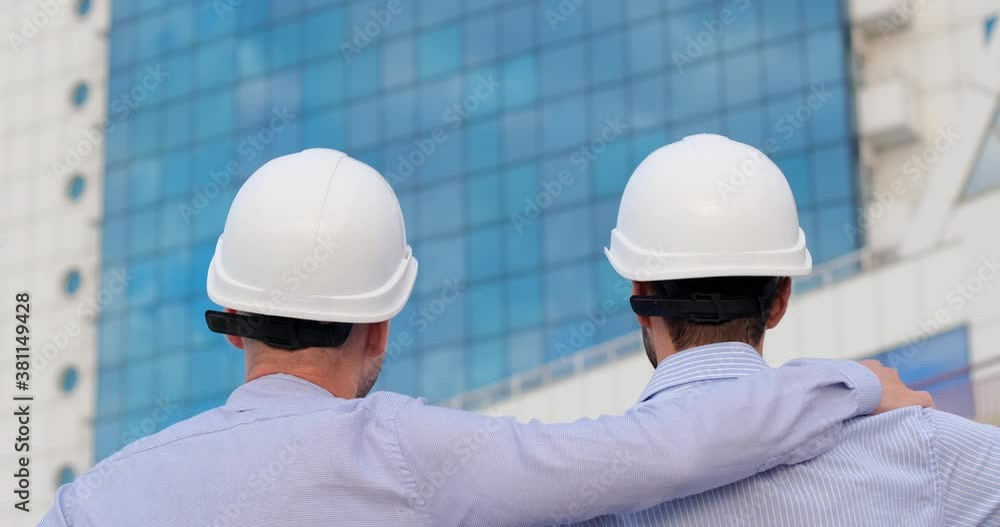 Two men, standing facing a building with blue windows, put on white protective helmets, an engineer hugs a colleague by the shoulder. Two men, standing facing a building with blue windows, put on white protective helmets, an engineer hugs a colleague by the shoulder.