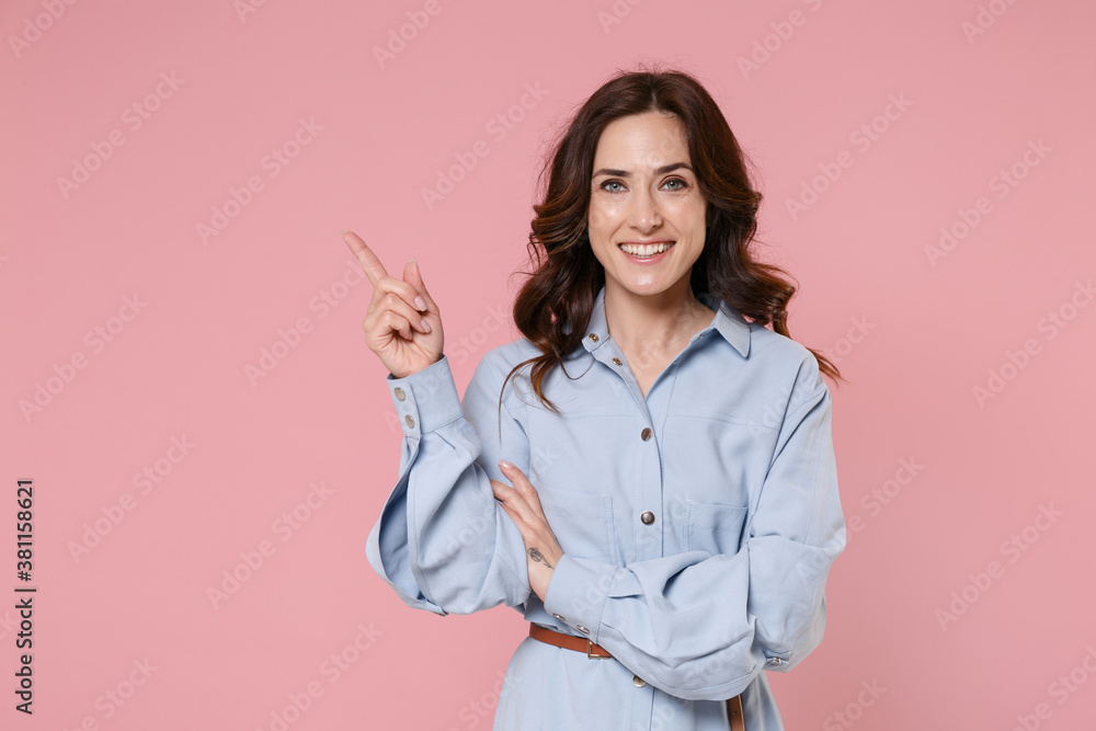 Smiling beautiful young brunette woman 20s in casual blue shirt dress posing standing pointing index finger aside up on mock up copy space isolated on pastel pink colour background studio portrait. Smiling beautiful young brunette woman 20s in casual blue shirt dress posing standing pointing index finger aside up on mock up copy space isolated on pastel pink colour background studio portrait.