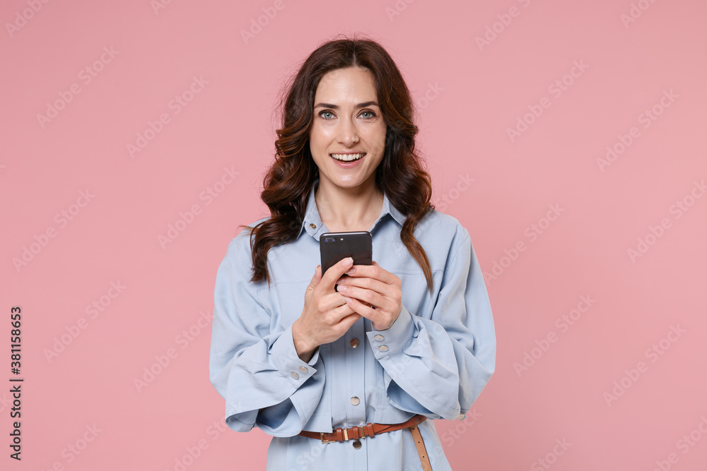 Smiling young brunette woman 20s wearing casual blue shirt dress posing standing using mobile cell phone typing sms message looking camera isolated on pastel pink colour background, studio portrait. Smiling young brunette woman 20s wearing casual blue shirt dress posing standing using mobile cell phone typing sms message looking camera isolated on pastel pink colour background, studio portrait.