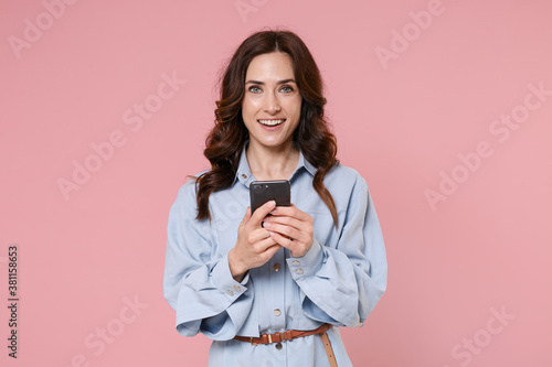 Smiling young brunette woman 20s wearing casual blue shirt dress posing standing using mobile cell phone typing sms message looking camera isolated on pastel pink colour background, studio portrait.